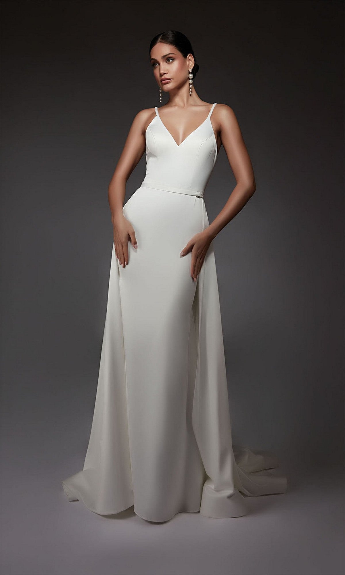 A woman stands against a dark background wearing the Alyce 7090 Long Formal Dress, a sleeveless, floor-length satin gown with a V-neckline that exudes elegance and timeless style.